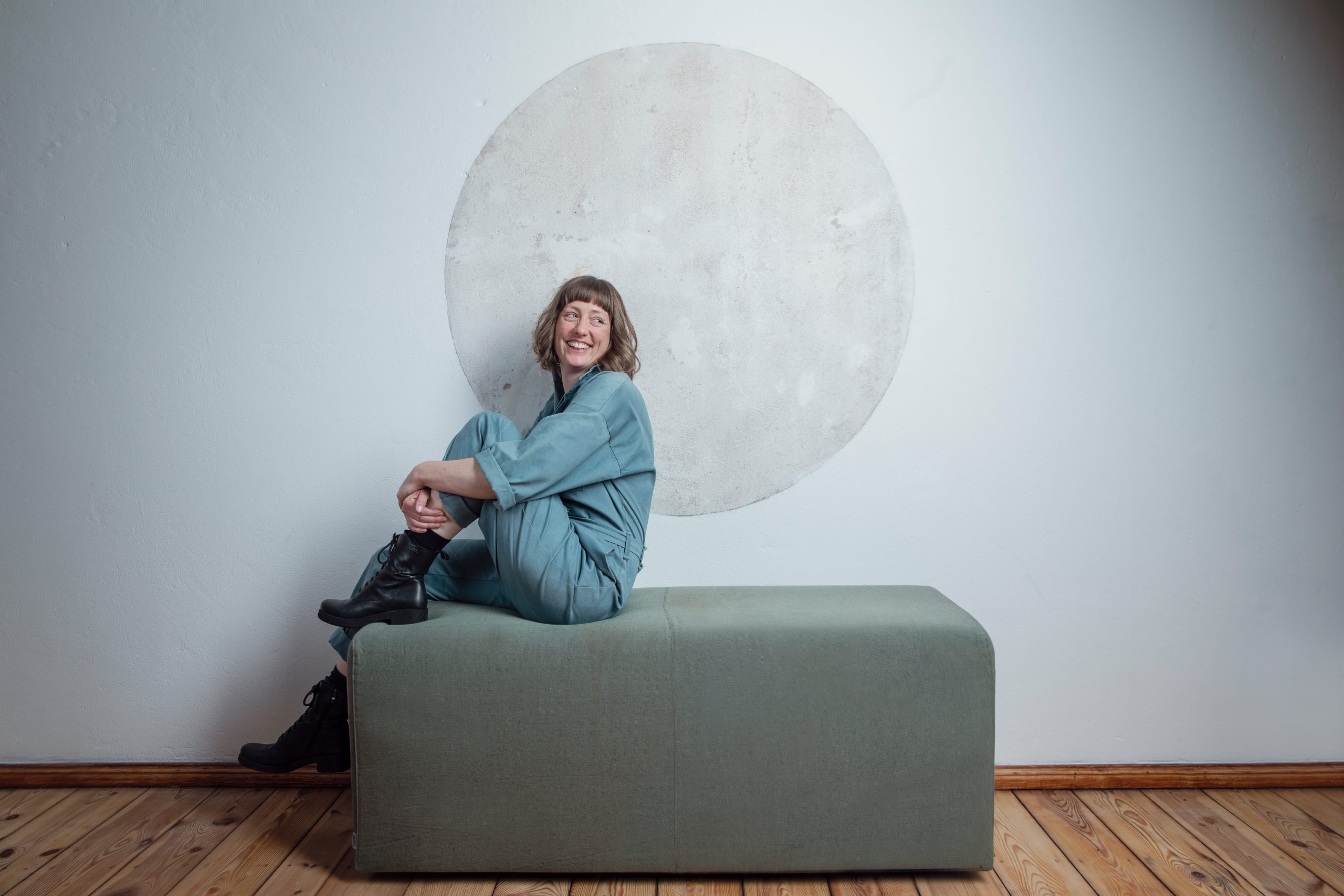 Ein Künstlerinnen-Portrait der Musikerin Teresa Bergman. Sie sitzt an der Seite eines Hockers und scheint jemanden neben der Kamera anzulachen