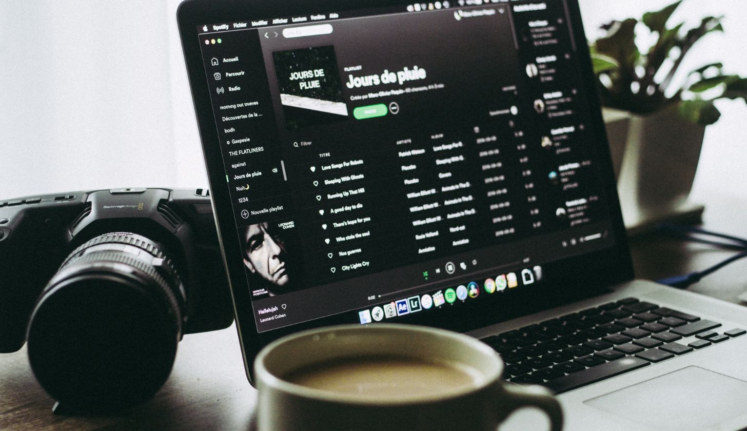 Auf einem Laptop ist Spotify geöffnet, daneben liegt eine Kamera und davor steht eine Kaffetasse
